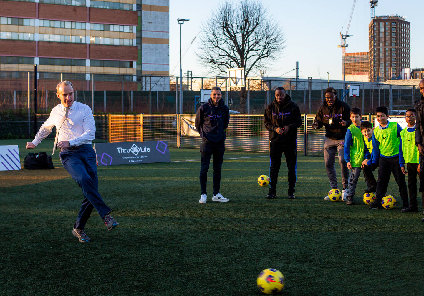 Deputy Prime Minister Dominic Raab in a shirt and tie kicks a ball as a group of coaches and children watch.