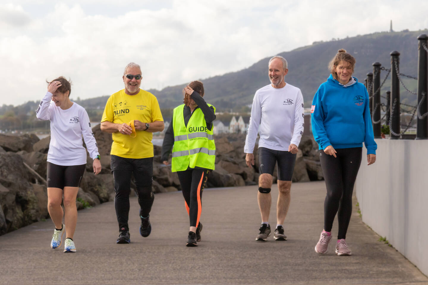 Man with a visual impairment running alongside his guide runner and other runners by the seaside.