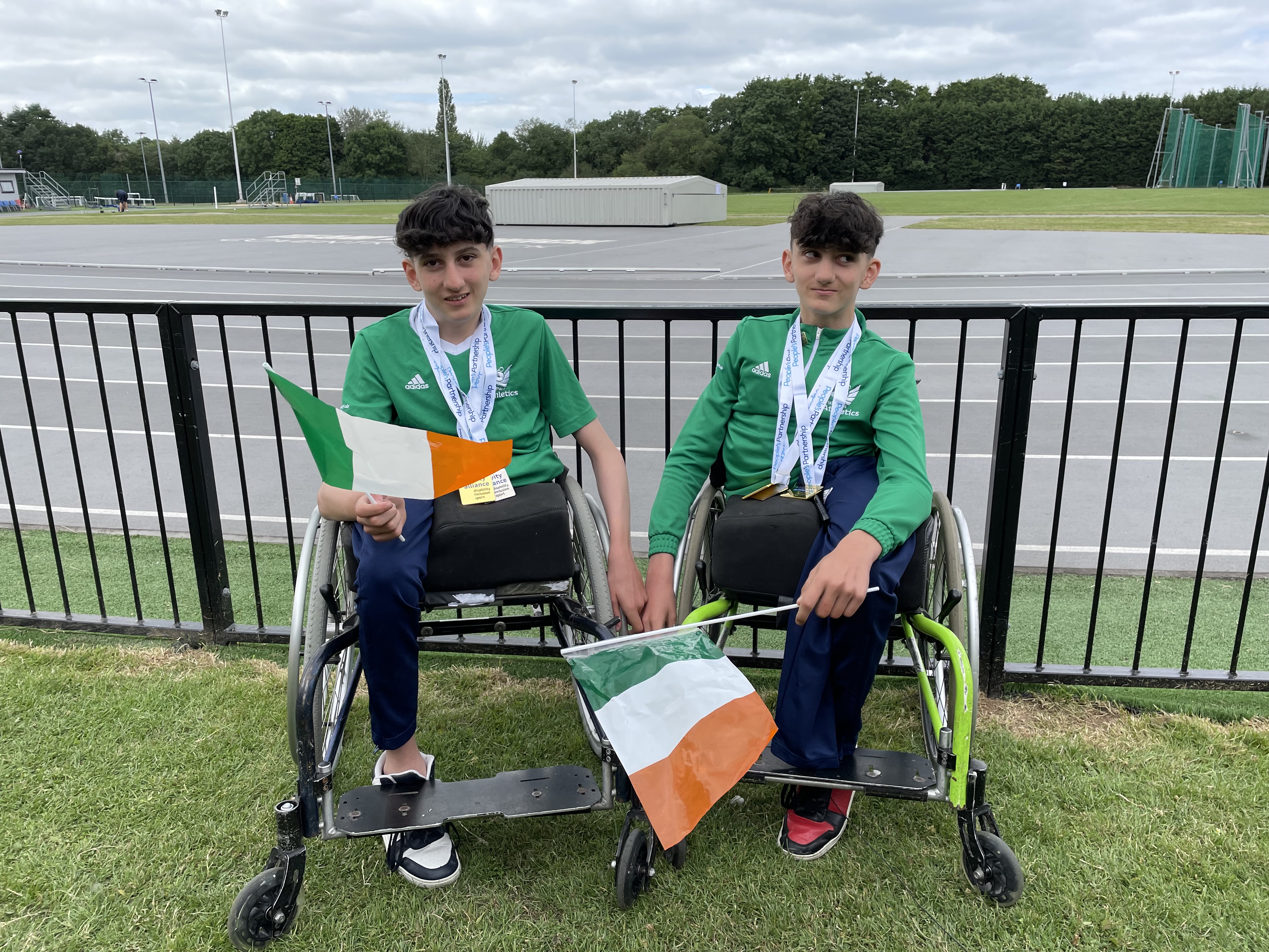 Two young boys in wheelchairs wave Irish flags in front of an athletics track.