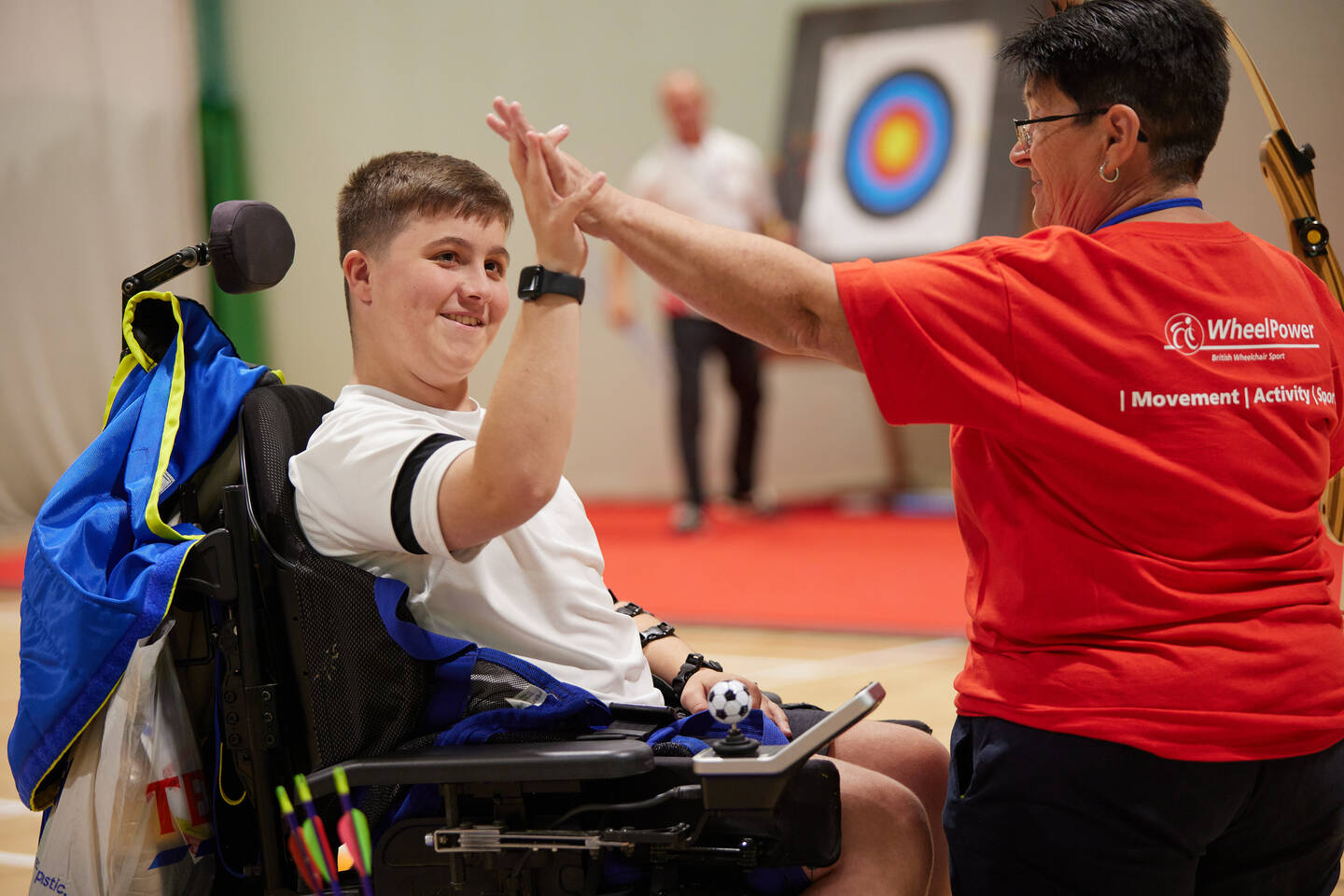 A young man in an electric wheelchair is taking part in the Wheelpower Junior Games. He is being 'high-fived' by a coach.