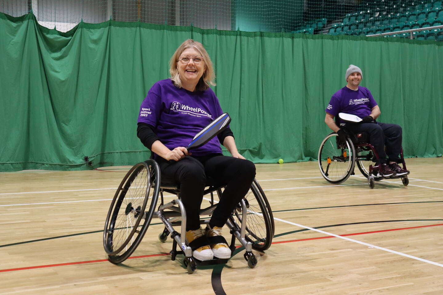 A woman in a wheelchair smiles holding a tennis racket.