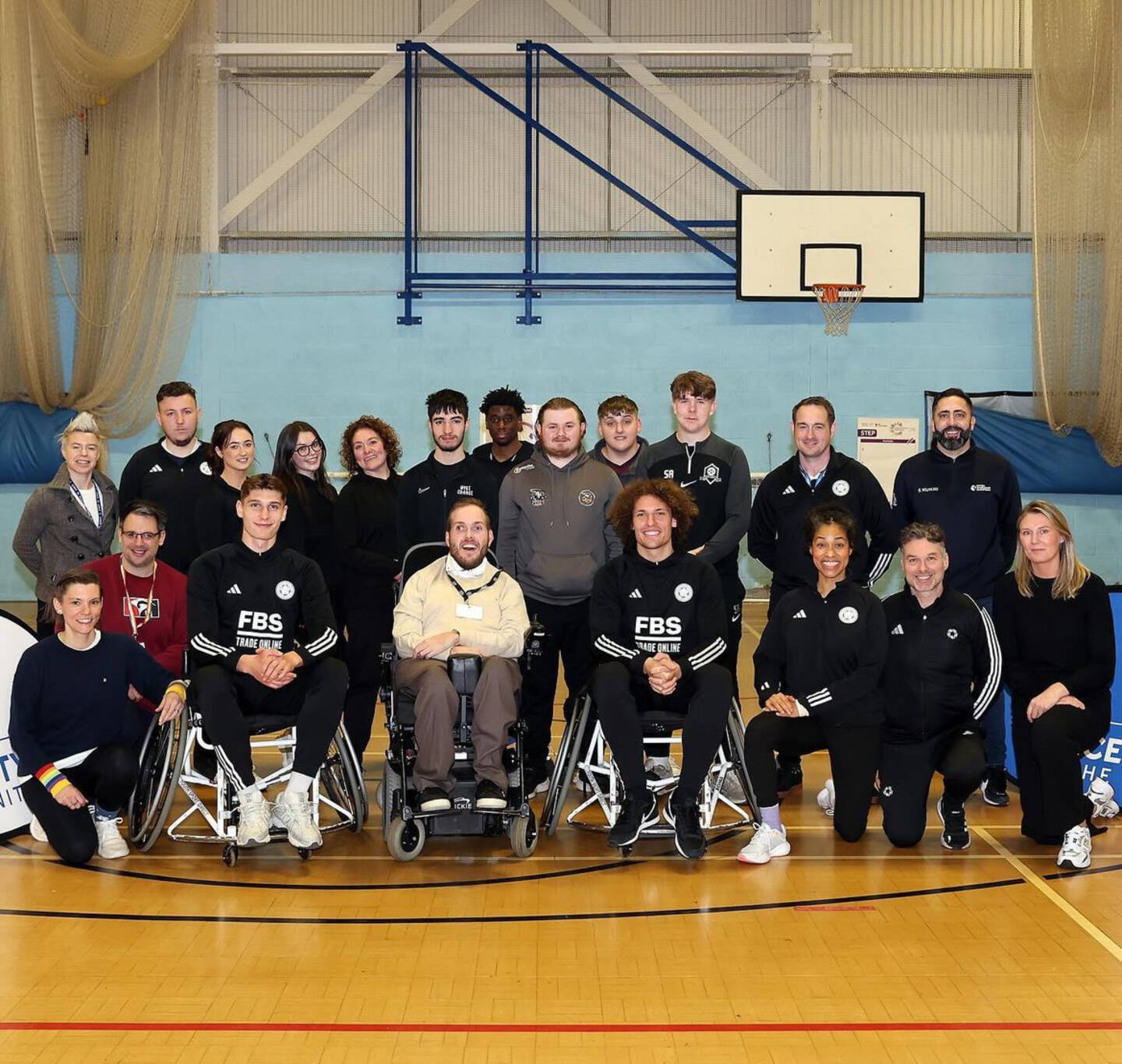 A group of people stand in a sports hall. Three people at the front of the group, including footballers Wout Faes and Ben Nelson, are using wheelchairs.