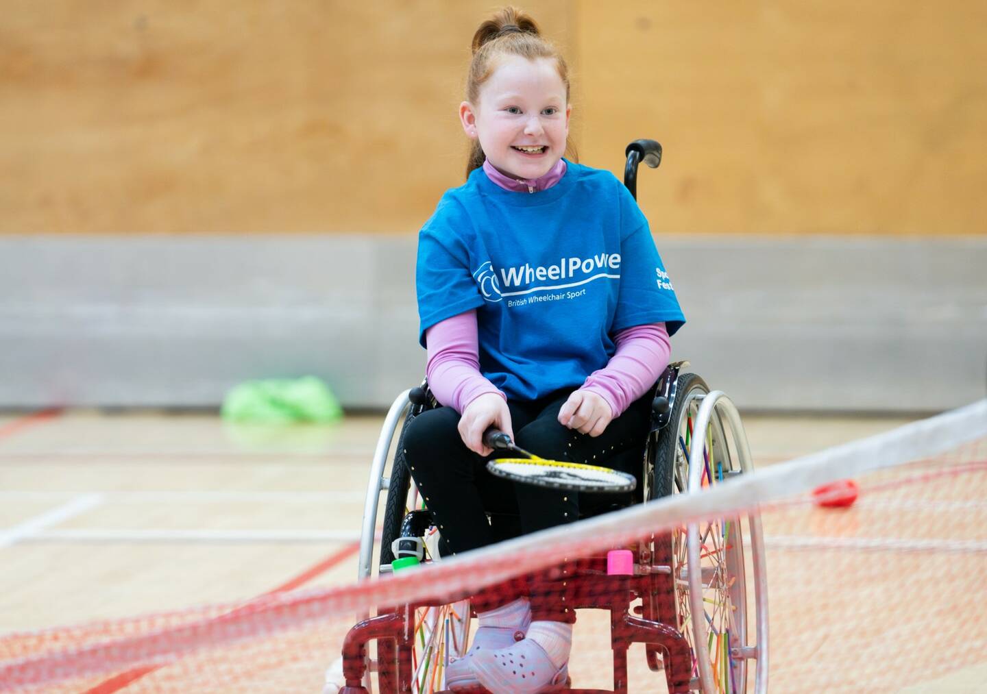 A young girl in a wheelchair is enjoying taking part in a game of badminton in a sports hall. She is wearing a blue 'Wheelpower' t-shirt.