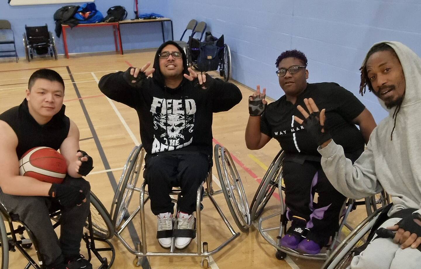 Group of disabled and non-disabled people posing for a picture with a basketball in sports wheelchairs in a sport hall.